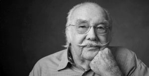 The black-and-white image shows a close-up portrait of William Sullivan. He is wearing glasses and has a distinctive mustache. His expression is thoughtful, with one hand resting under his chin. The background is simple and dark, drawing attention to his face. The portrait captures a sense of wisdom and character.