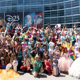 An image showing fans from a previous D23 Cosplay Meet-Up. Various fans of Disney Princesses franchises stand and kneel in front of the camera to showcase their cosplay creations in front of the Anaheim Convention Center. On the building is a billboard with the writing “D23 The Ultimate Disney Fan Event.”