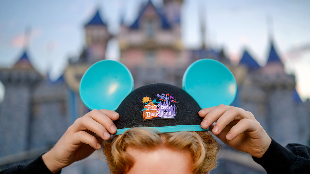 The image shows a close-up of a person adjusting a Mickey Mouse ear hat. The hat is black with blue ears and a blue trim, featuring an embroidered design with "D23 Day at Disneyland Resort" in colorful text alongside images of iconic Disneyland symbols, including the Sleeping Beauty Castle and fireworks. The background is blurred, but it is clear that the person is standing in front of Sleeping Beauty Castle at Disneyland.