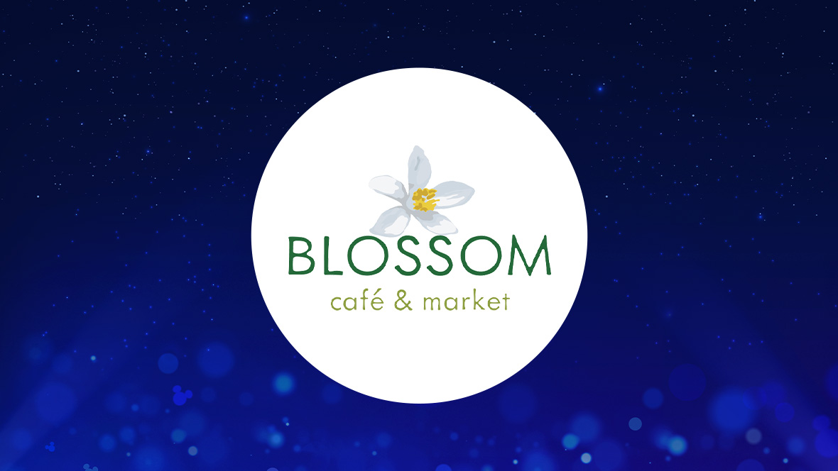 The image features a white circle centered on a deep blue background with subtle light effects and sparkling points. Inside the circle, the text "BLOSSOM café & market" is displayed. Above the text is a delicate illustration of a white blossom with a yellow center. The word "BLOSSOM" is written in a green, uppercase font, while "café & market" is in a smaller, lowercase font in light green.