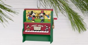 A colorful Christmas ornament featuring a vintage-style piano with Disney characters. The piano has a red and green color scheme with Mickey Mouse, Minnie Mouse, Goofy, and Donald Duck depicted in a lively musical scene on the top section. Below, the piano keys and a small illustration of Mickey Mouse and Minnie Mouse are visible, along with the text 'Mickey Mouse' and musical notes on a sheet. The ornament is hanging from a Christmas tree branch in the background.