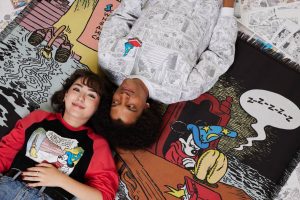 Two people lying down on a colorful blanket featuring illustrations from 'The Sorcerer's Apprentice' with Mickey Mouse. The person on the left has short dark hair, is smiling, and wearing a red and black shirt with a comic strip image of Mickey Mouse. The person on the right has curly hair and is wearing a light-colored shirt with comic strip designs. They are both surrounded by comic strip pages.