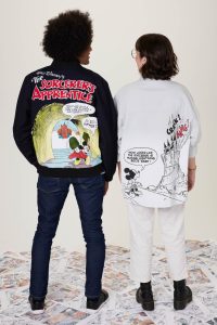 Two people standing with their backs to the camera. The person on the left has curly hair and is wearing a black jacket with a colorful 'The Sorcerer's Apprentice' design featuring Mickey Mouse. The person on the right has short dark hair and is wearing a light-colored shirt with a black and white comic strip design, including Mickey Mouse and a castle with 'CRACK! BANG!' text. Both are standing on a floor covered with comic strip pages.