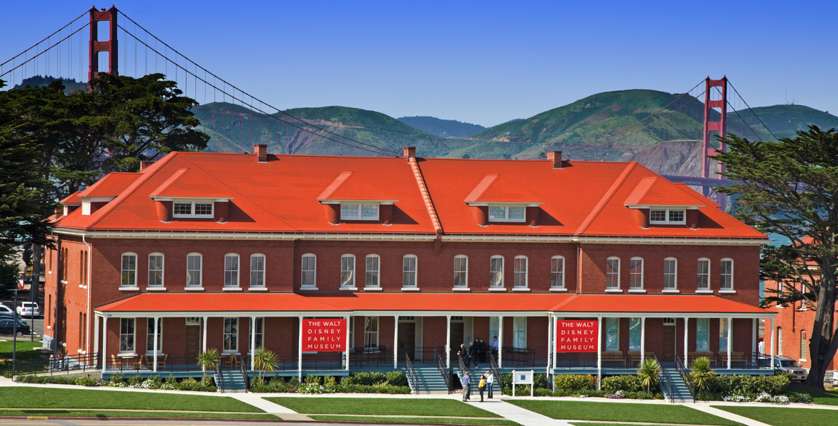 Exterior view of The Walt Disney Family Museum with the Golden Gate Bridge visible in the background. The museum is housed in a red-brick building with a red roof, and features a sign that reads 'The Walt Disney Family Museum' on the front facade. The setting includes a manicured lawn and a few visitors near the entrance.