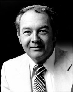 The black-and-white image is a portrait of William Sullivan. He is wearing a light-colored suit with a white shirt and a striped tie. His hair is neatly combed, and he has a calm and composed expression on his face. The background is dark, which highlights his features and attire. The portrait captures a professional and dignified appearance.