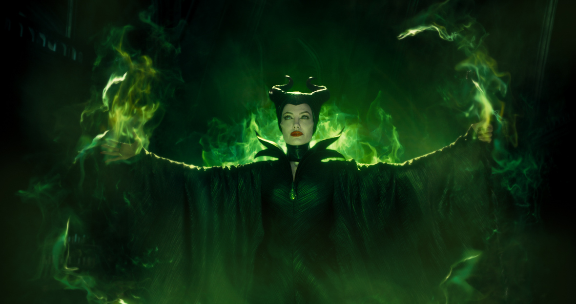 In a scene from Maleficent: Mistress of Evil, Maleficent (Angelina Jolie) wears a black turtleneck cloak, her arms spread to the side. She wears a horn headdress and wears dark red lipstick, while neon green mist surrounds her body.