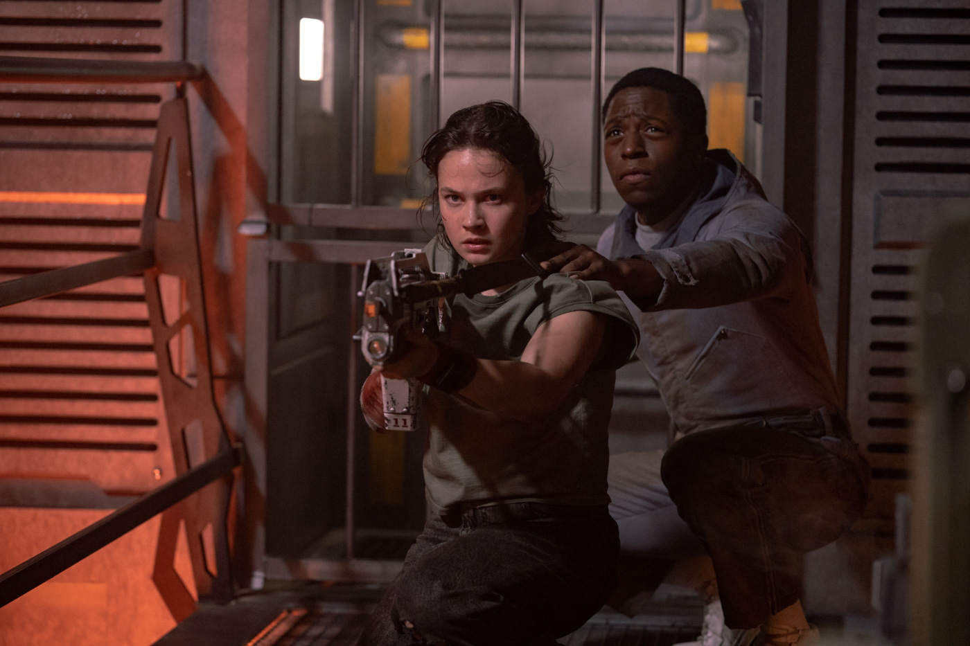 Rain Carradine (Cailee Spaeny) aims a blaster as Andy (David Jonsson) places his left hand on her left shoulder. They are crouching near a door on an abandoned spaceship.