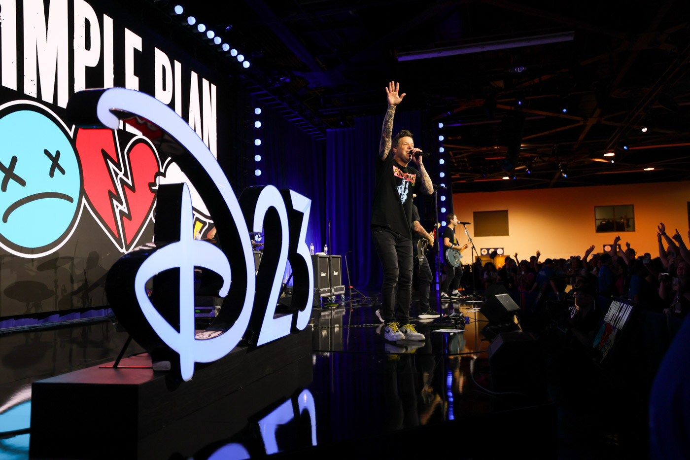 On a stage reading “Simple Plan” with a broken heart tattoo and an x-eyed smiley face and “D23,” Pierre Bouvier sings and Sebastien Lefebvre is playing the electrical guitar. To their right is a crowd of fans, dancing and singing along. 