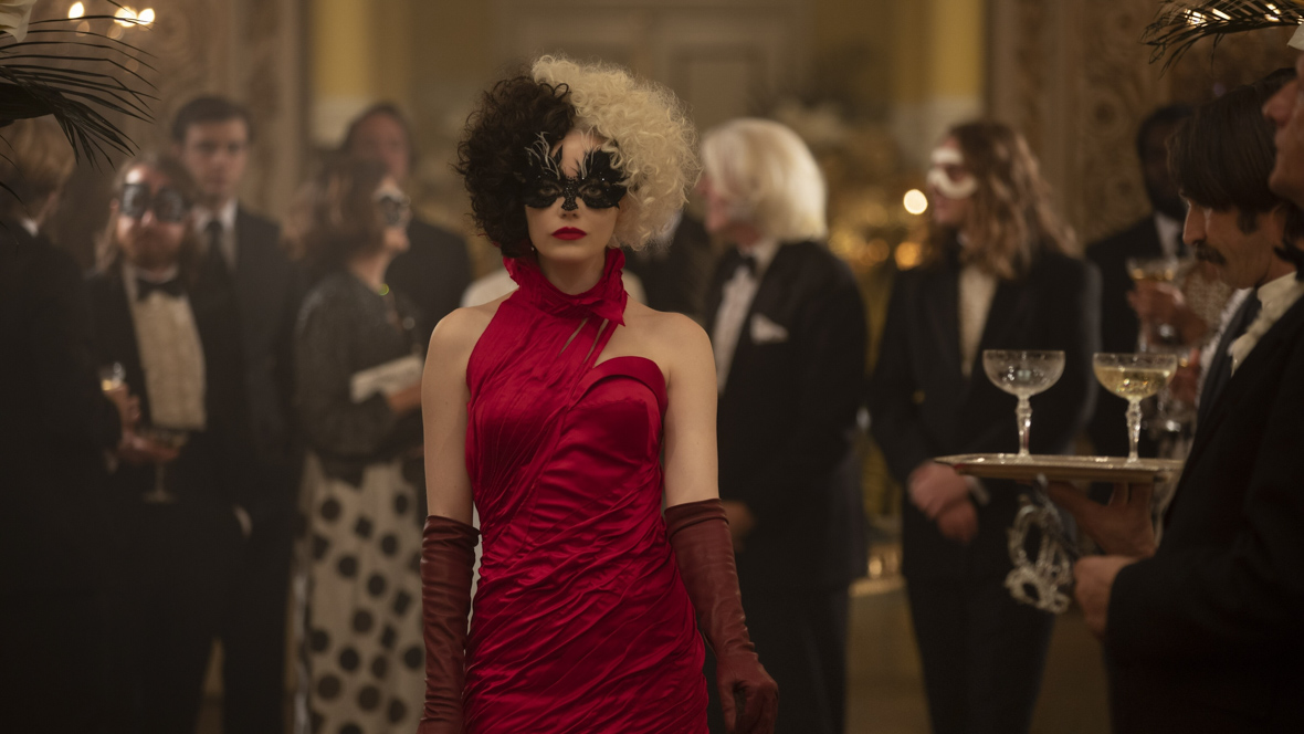 In a scene from Cruella, Emma Stone as Cruella walks through a black-tie party wearing a crimson red, one-shoulder, high-neck dress paired with burgundy gloves. Her hair is half white, half black, and she wears a black mask covering the upper part of her face. A server to her right holds a tray with drinks, while party guests in formal attire converse in the background.