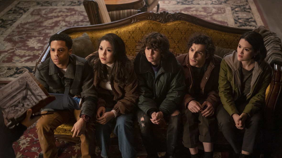 In a scene from Goosebumps (2023), from left to right, Isaiah (Zack Morris), Isabella (Ana Yi Puig), James (Miles McKenna), Lucas (Will Price), and Margot Stokes (Isa Briones) are in a living room, sitting on a mustard-brown couch, all gazing upward with eyes completely black, indicating possession by a mysterious force. In the lower left corner, Mr. Bratt (Justin Long) holds a scrapbook. The background features two chairs, a wooden table, and a blue and red patterned carpet. 