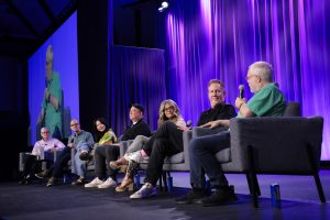 From left to right, Don Peri, Peter Docter, Domee Shi, Peter Sohn, Jennifer Lee, Jared Bush, and Ron Clements are sitting on grey sofa chairs on a stage. The background includes blue stage curtains and a screen showing a live video of Ron Clements speaking into a microphone.