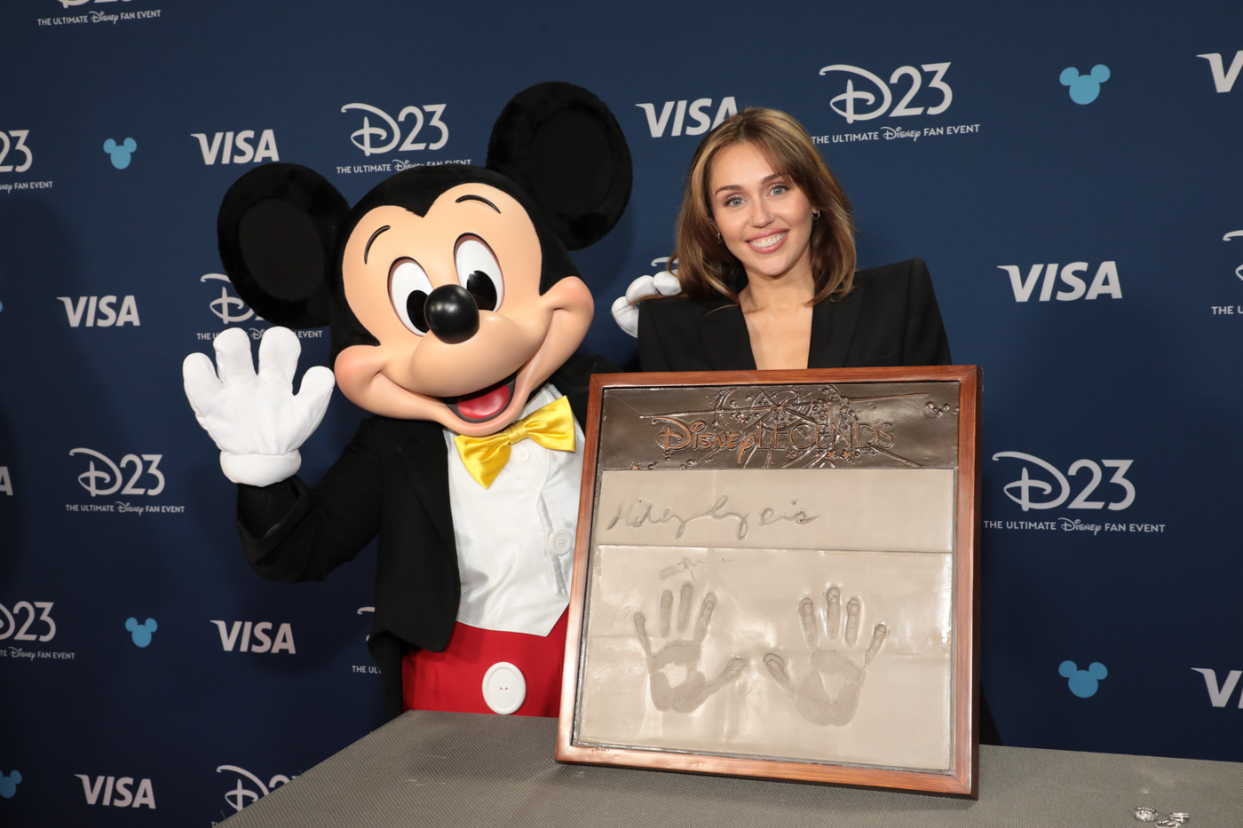 At the Disney Legends Handprint Ceremony, Disney Legend Miley Cyrus (right) poses with her handprint alongside Mickey Mouse (left). Cyrus is dressed in a black blazer, while Mickey waves at the camera. The backdrop features graphics reading “D23 The Ultimate Disney Fan Event,” “VISA,” and small blue Mickey Mouse symbols.