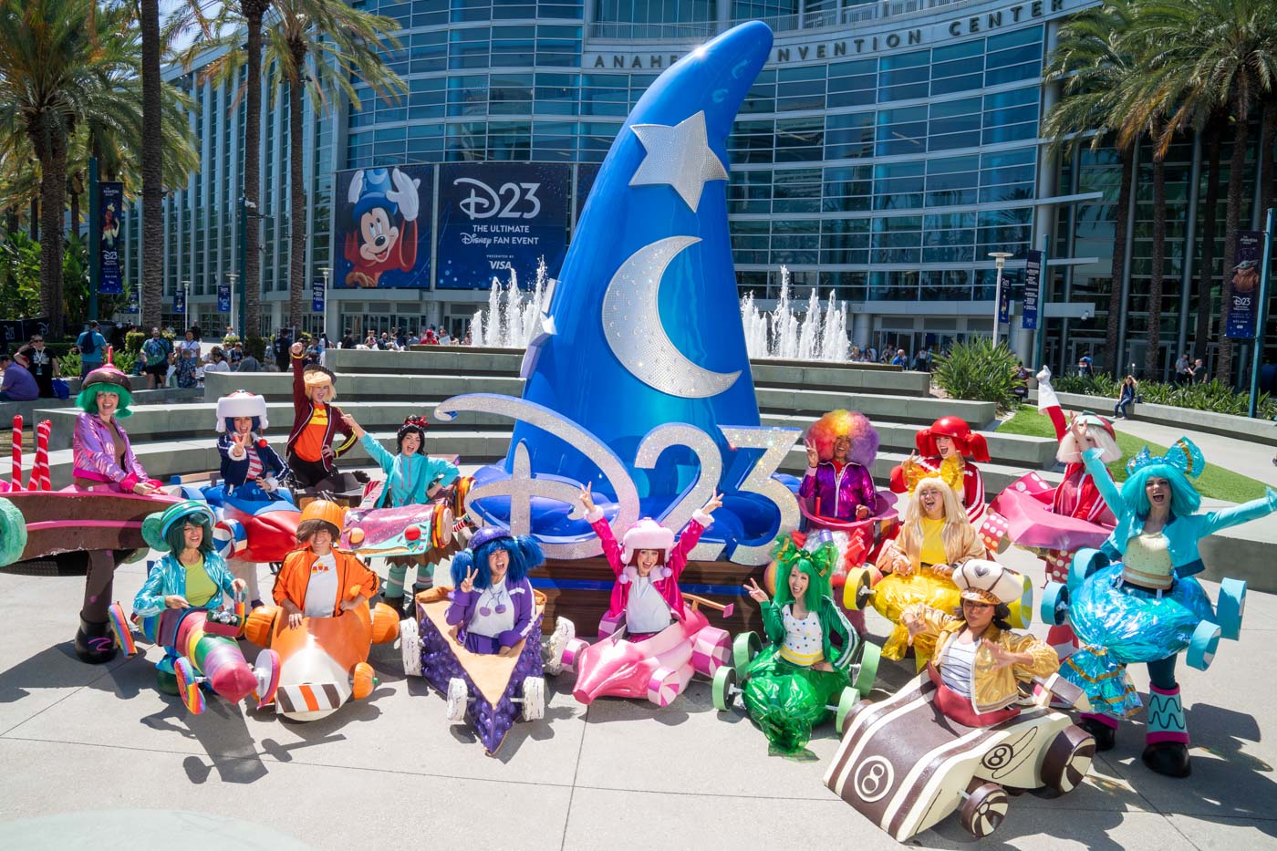 Fifteen fans dressed as Sugar Rush racers pose around a Sorcerer Mickey hat with a sparkling “D23” sign outside the Anaheim Convention Center. Behind them, a fountain and a poster reading “D23 The Ultimate Disney Fan Event Presented by VISA” are visible on the convention center building.