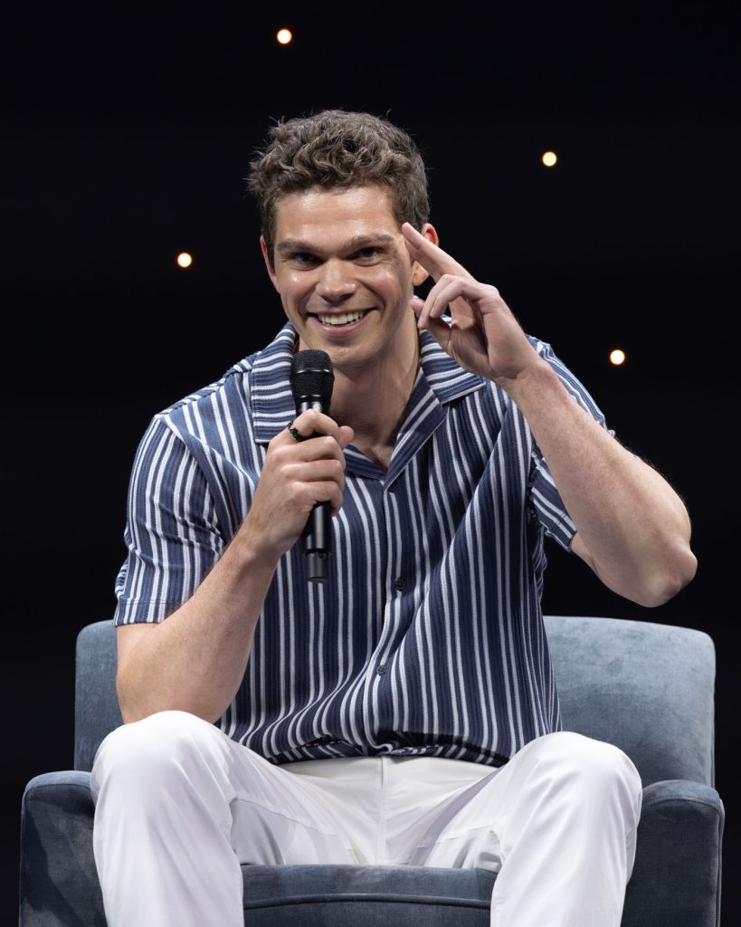 Daniel Diemer sits on a gray sofa chair during D23’s Percy Jackson... panel, wearing a blue and white striped short-sleeve shirt and white pants. He holds a microphone in his right hand, smiling warmly.