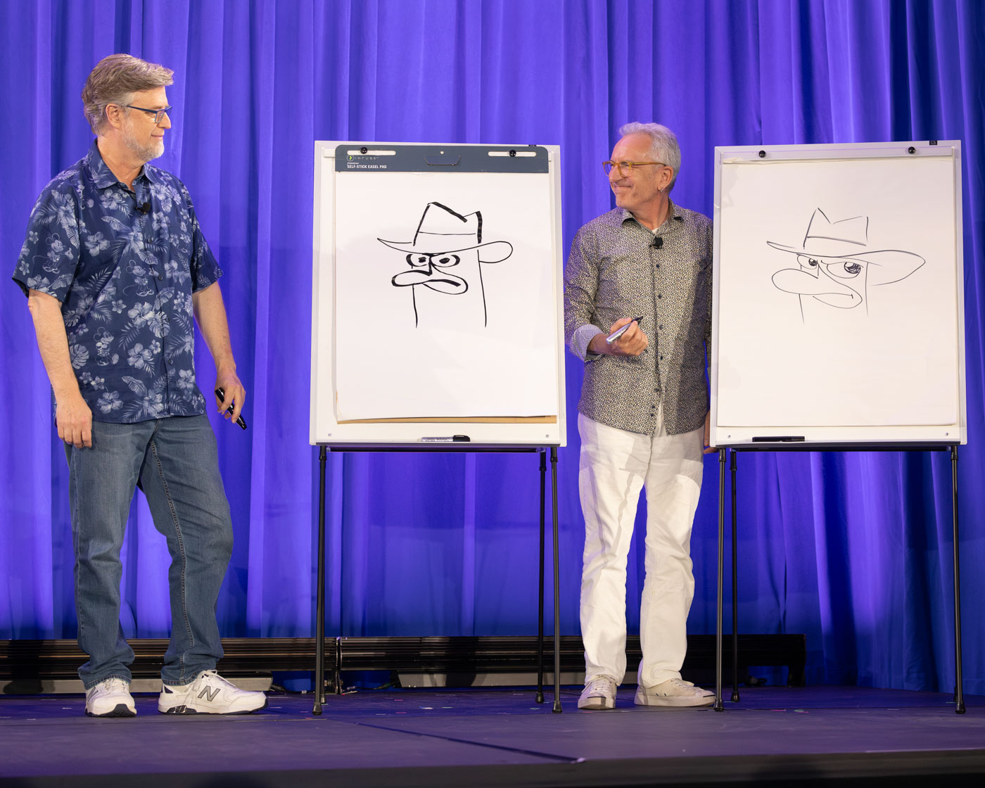 Dan Povenmire (left) and Jeff “Swampy” Marsh (right) are on a stage in front of a blue curtain at D23 2024, each standing next to an easel with a large drawing pad. Both men are smiling as they each hold a marker, having just drawn Perry the Platypus, a platypus detective from the show Phineas and Ferb. 