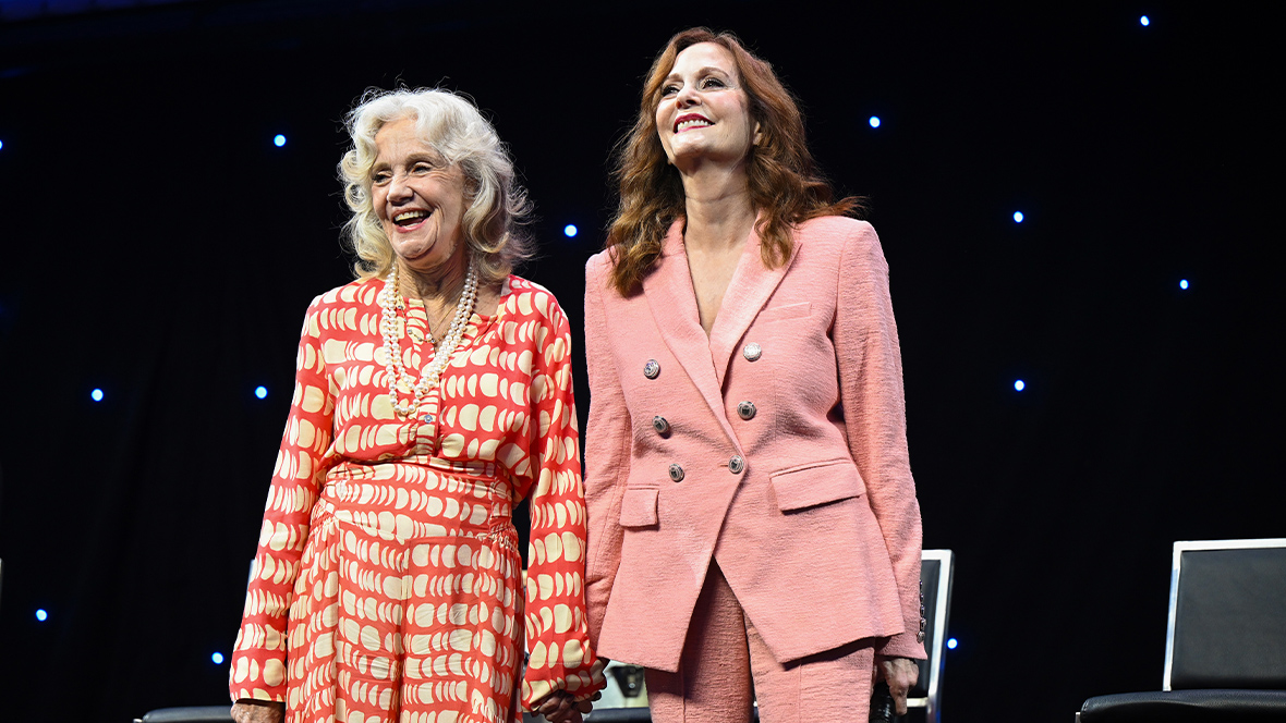 Disney Legend Hayley Mills (left) and Lesley Ann Warren (right) are on a stage holding hands. Mills is wearing a long sleeve dress with red and white prints, holding a microphone in her right hand. Warren is wearing a pink blazer with silver buttons and pants, holding a microphone in her left hand. 