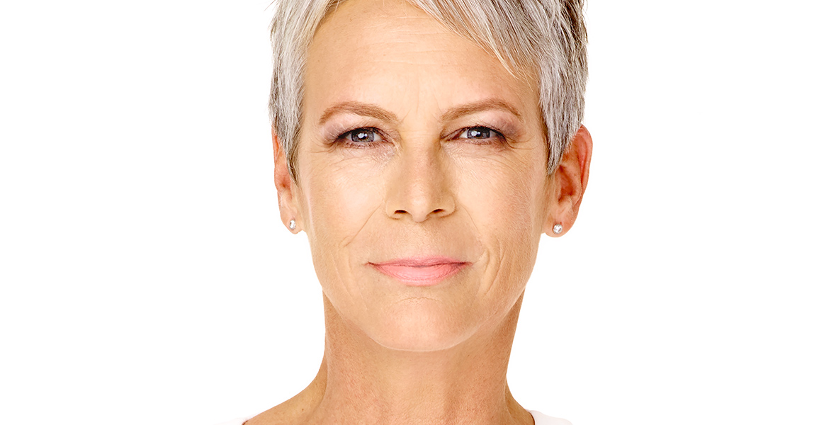 Disney Legend Jamie Lee Curtis sports short silver hair, wearing subtle makeup and small stud earrings, looking directly at the camera with a neutral expression against a plain white background.