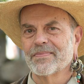 The image features Disney Legend Joe Rohde. He is seen wearing a straw hat and has a distinguished beard. Joe Rohde is also notable for his unique earring, which includes multiple charms and beads.