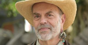 The image features Disney Legend Joe Rohde. He is seen wearing a straw hat and has a distinguished beard. Joe Rohde is also notable for his unique earring, which includes multiple charms and beads.