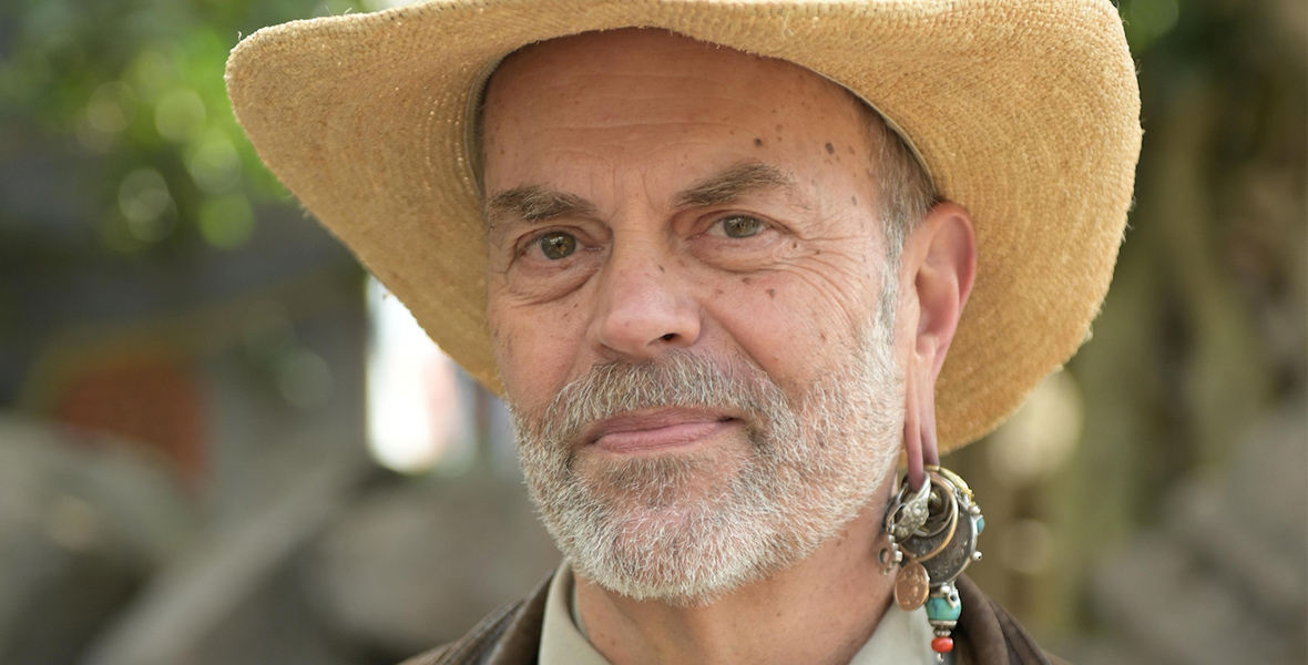 The image features Disney Legend Joe Rohde. He is seen wearing a straw hat and has a distinguished beard. Joe Rohde is also notable for his unique earring, which includes multiple charms and beads.