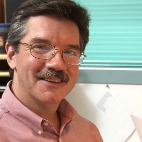 Disney Legend Mark Henn sports glasses and a mustache, wearing a light pink shirt, smiling at the camera while seated at a desk. The background includes a shelf with various items and a window with blinds.