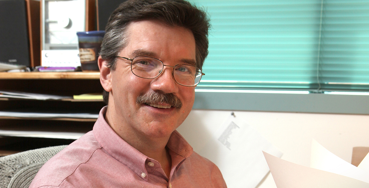 Disney Legend Mark Henn sports glasses and a mustache, wearing a light pink shirt, smiling at the camera while seated at a desk. The background includes a shelf with various items and a window with blinds.