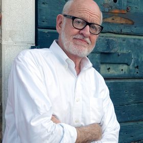 Disney Legend Frank Oz sports a white beard and glasses, wearing a white button-down shirt, standing with his arms crossed against a backdrop of a brick wall and a weathered wooden door.