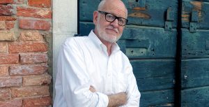 Disney Legend Frank Oz sports a white beard and glasses, wearing a white button-down shirt, standing with his arms crossed against a backdrop of a brick wall and a weathered wooden door.