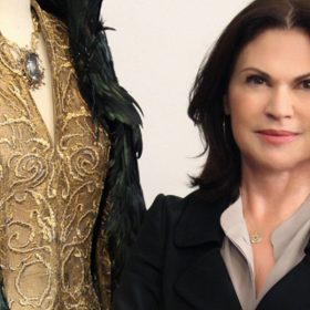 Disney Legend Colleen Atwood sports dark brown hair, wearing a black blazer and a light-colored blouse, stands next to an ornate gold and green feathered costume.