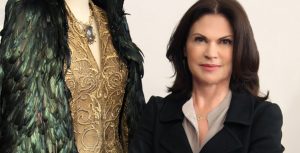 Disney Legend Colleen Atwood sports dark brown hair, wearing a black blazer and a light-colored blouse, stands next to an ornate gold and green feathered costume.