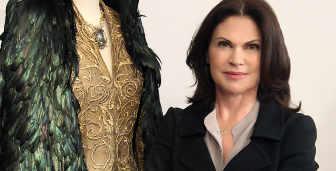 Disney Legend Colleen Atwood sports dark brown hair, wearing a black blazer and a light-colored blouse, stands next to an ornate gold and green feathered costume.