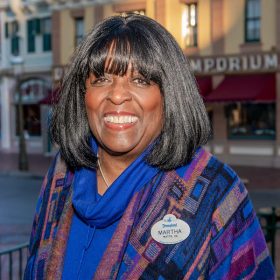 Disney Legend Martha Blanding sports shoulder-length dark hair, wearing a blue and patterned shawl with a Disneyland name tag, smiles while standing in front of a background of shops and buildings.