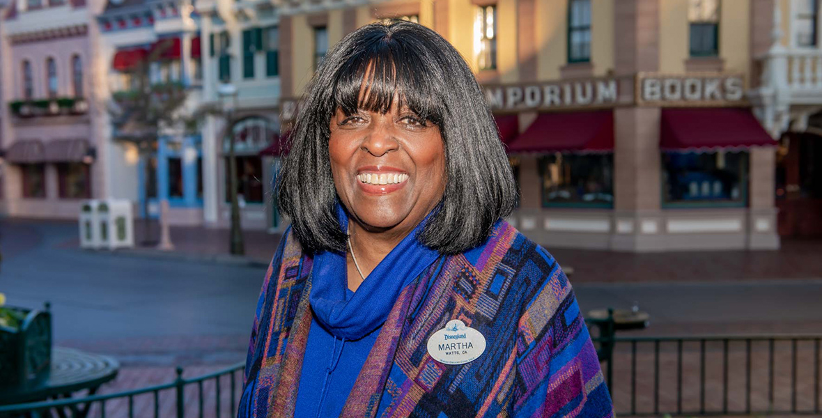 Disney Legend Martha Blanding sports shoulder-length dark hair, wearing a blue and patterned shawl with a Disneyland name tag, smiles while standing in front of a background of shops and buildings.