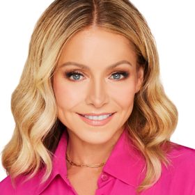 Disney Legend Kelly Ripa sports shoulder-length blonde hair, wearing a bright pink shirt, smiles at the camera with a white background.