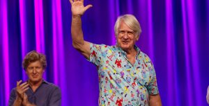 Disney Legend Bill Farmer, the official voice of Goofy, waves to the crowd as he joins a panel about the making of The Goofy Movie at D23: The Ultimate Fan Event. Steve Moore is seen applauding behind him.