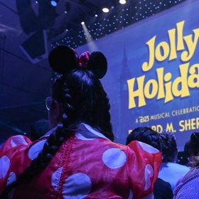 Seen from the perspective of the audience is part of the stage for the “Jolly Holiday: A D23 Musical Celebration of Richard M. Sherman” during D23: The Ultimate Disney Fan Event 2024. The logo for the show can be seen on the stage’s large screen backdrop, and in the foreground is an audience member wearing Minnie Mouse ears and a red polka-dot dress.