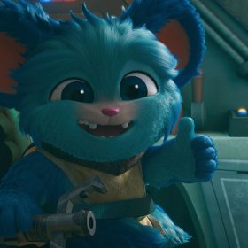 In a scene from the “Star Wars: Young Jedi Adventures,” Nubs (a blue, furry member Poobian species voiced by Dee Bradley Baker), is in a seat inside a blue spacecraft. He is signing a “thumbs up” with his left hand, smiling at the camera. He wears mustard-brown Jedi apparel.