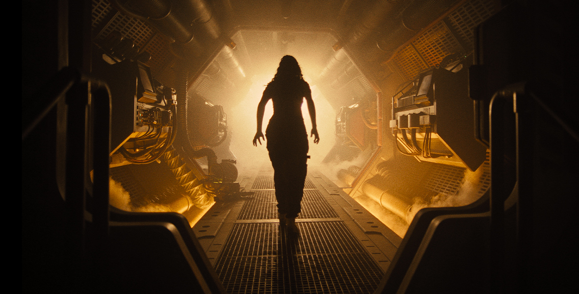 Kay (Isabela Merced) walks through a corridor on a spaceship. Her silhouette is illuminated by a bright orange light.