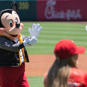 Mickey Mouse is seen clapping his hands while standing on a baseball field, dressed in a casual outfit with a black vest and red pants. In the background, a man wearing a white Disney baseball jersey and a red cap walks on the field, and a person in the foreground wearing a red cap faces Mickey.