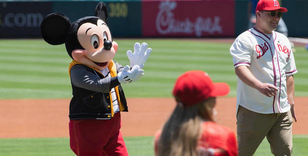 Mickey Mouse is seen clapping his hands while standing on a baseball field, dressed in a casual outfit with a black vest and red pants. In the background, a man wearing a white Disney baseball jersey and a red cap walks on the field, and a person in the foreground wearing a red cap faces Mickey.