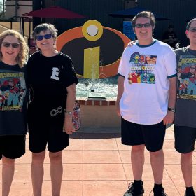 Four people are posing in front of an outdoor fountain, which features the iconic "Incredibles" logo. The group is wearing themed T-shirts, with some sporting "The Incredibles" characters and Pixar designs. T