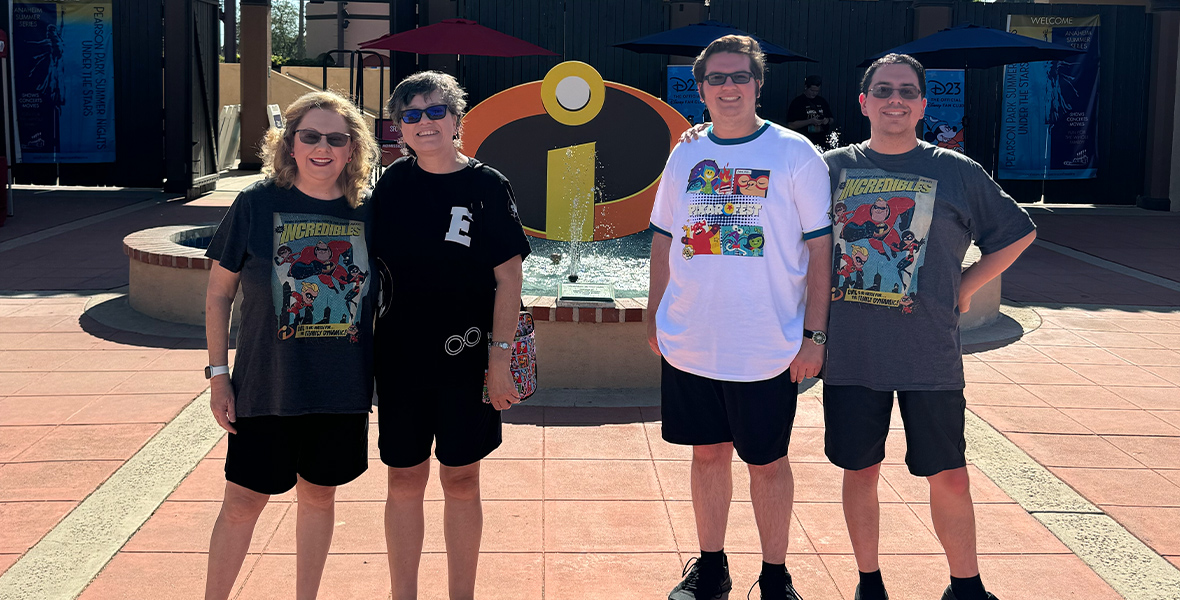 Four people are posing in front of an outdoor fountain, which features the iconic "Incredibles" logo. The group is wearing themed T-shirts, with some sporting "The Incredibles" characters and Pixar designs. T