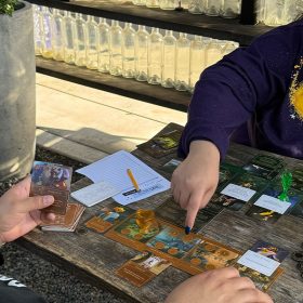 Two people are seated at a wooden table outdoors, engaged in a card game. The person on the right is smiling and pointing at the game board, while the person on the left is holding a card and focused on the gameplay. The table is filled with various game pieces, cards, and coins, with a notepad and pencil nearby.