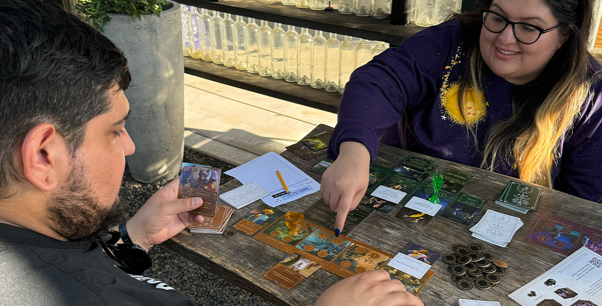 Two people are seated at a wooden table outdoors, engaged in a card game. The person on the right is smiling and pointing at the game board, while the person on the left is holding a card and focused on the gameplay. The table is filled with various game pieces, cards, and coins, with a notepad and pencil nearby.