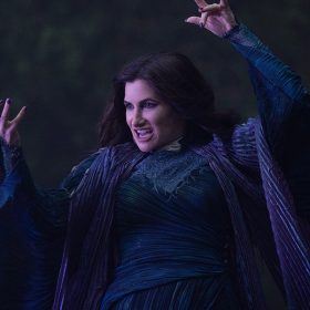 Agatha Harkness (Kathryn Hahn) from Marvel Television’s Agatha All Along wears a long-sleeved, pleated dark blue dress with dark purple fabric draped around her neck. She is pictured using her powers with slightly curled hands and purple-painted nails, speaking against a blurred, dark-toned background.