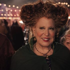 In a scene from Hocus Pocus 2 (from left to right), Mary Sanderson (Kathy Najimy), Winifred Sanderson (Disney Legend Bette Midler), and Sarah Sanderson (Sarah Jessica Parker) are standing, looking slightly to their right. Mary and Sarah are wearing burgundy cloaks, and Winifred wears a green cloak. In the background are outdoor string lights.