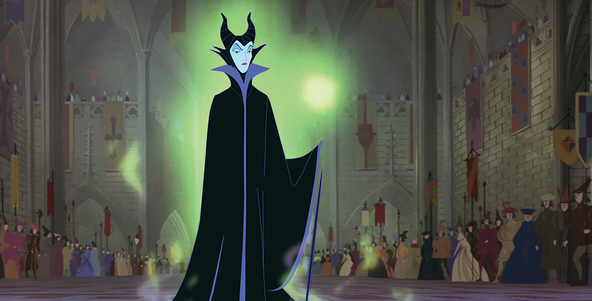 In a scene from Sleeping Beauty (1959), Maleficent (voiced by Eleanor Audley) is standing in a grand, Gothic-inspired Hall. She is wearing a black robe and horned headdress, with a high collar and a glowing green aura around her. The room is vast, with high ceilings, stone walls, and hanging banners. In the background, people are rushing toward an exit.