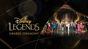 A group of Disney Legends honorees standing on stage at the Disney Legends Awards Ceremony. The stage is adorned with elegant gold and black decorations, with the Disney Legends logo prominently displayed above the group. Mickey Mouse and Minnie Mouse, dressed in their classic outfits, are standing at either end of the lineup of honorees.