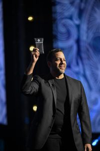 David Blaine stands on a stage at the Honda Center during D23 2024, holding out a deck of cards in a black plastic case labeled “DO NOT OPEN.” He is dressed in a black suit and t-shirt, with blue projections illuminating the background.