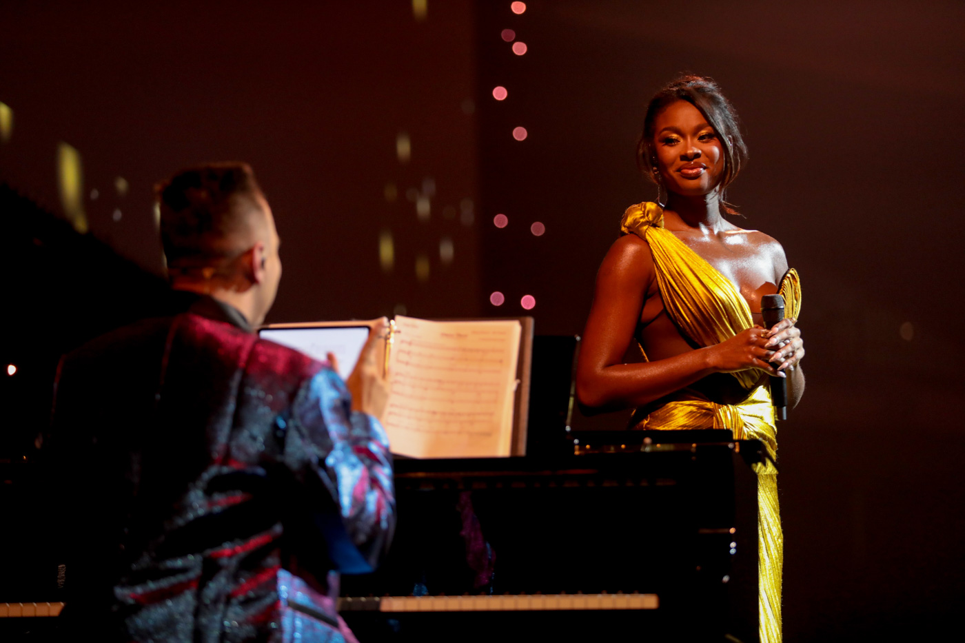 Coco Jones (right) is wearing golden one-shoulder gown stands on stage during the Disney Princess concert at D23: The Ultimate Disney Fan Event 2024, holding a microphone. To her right is a musician; in front of her, a man, dressed in a shiny, dark-colored suit, is seated at a piano, playing music from a sheet on the stand. The stage is softly lit with a backdrop of subtle, glowing orbs. 